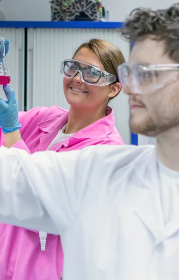 Female student in pink lab coat holding up flask while male student focuses on pipette Female student in pink lab coat holding up flask while male student focuses on pipette